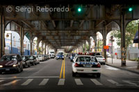 Típico paisaje de Queens, con el metro elevado por encima de la carretera y los coches de policía vigilando la zona. Este barrio debe el nombre a la reina Catalina de Braganza, esposa de Carlos II de Inglaterra. Esto sucedió a finales del siglo XVII, los primeros habitantes que se instalaron aquí fueron modestos granjeros. El barrio experimentó un boom demográfico con la llegada del tren en 1910. En los años 1920 y 1930, Queens vivió una época de esplendor con la instalación de los estudios Paramount que enseguida pasaron a llamarse Astoria Movie Studio y se convirtió en la capital del cine en la costa este. Aquí es donde Rodolfo Valentino, los hermanos Marx y muchos otros se pusieron por primera vez delante de una cámara. Lo malo fue la desaparición del cine mudo, que supuso también el fin de la hegemonía de los estudios. La población de Queens siguió aumentando y acogiendo cada vez más etnias que tenían un punto en común: la escasez de medios. Hoy es el barrio más multiétnico de Nueva York. Aquí las culturas cohabitan unas con otras, cada una con su propio ritmo. Al desembarcar en Queens, uno siente latir otro ritmo de vida, los edificios son más bajos, las aceras están más despejadas y la gente va con menos prisa. Durante mucho tiempo considerado como un barrio desheredado, hoy se abre al turismo, mientras conserva sus tesoros arquitectónicos y sus recuerdos. Aquí vivieron Jack Kerouac, Will Rogers, Heinrich Steinwerg, Billie Holiday, Ella Fitzgerald, Louis Amstrong, Dizzie Gillespie, Fats Waller y Count Basie, entre otros.