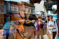 Juneau, Alaska, USA. People reflected in the showcase of Totem Pole Shop, Juneau Alaska. Indigenous crafts shop, Juneau, Alaska. Local Indian Art and Collectibles. Store offering local Indian art and collectibles. Title taken from label on matting. View of large and small totem poles in front of curio shop in Juneau, Alaska, with clock at left of large totem pole. Signs on building read: "Curios. Visitors welcome." and "The Nugget Shop." 