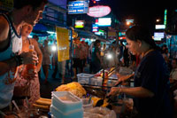 Bangkok. Khao San Road, puesto de fideos calle. Puesto de comida. Bangkok. Khaosan Road o Khao San Road es una calle corta en el centro de Bangkok, Tailandia. Es en la zona de Banglamphu (distrito de Phra Nakhon) cerca de 1 kilómetro (0,62 millas) al norte del Gran Palacio y el Wat Phra Kaew. "Khaosan" se traduce como "arroz blanco", un recordatorio de que en otros tiempos la calle era un importante mercado de arroz Bangkok. En los últimos 20 años, sin embargo, Khaosan Road se ha convertido en un famoso "gueto mochilero". Ofrece alojamiento económico, que van desde hoteles de estilo "colchón en una caja" a un precio razonable hoteles de 3 estrellas. En un ensayo sobre la cultura mochilero de Khaosan Road, Susan Orlean lo llamó "el lugar de desaparecer".