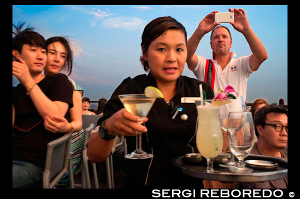 Cambrera amb un còctel a la mà. Banyan Tree terrat Vértigo i Moon Bar, Restaurant ,, Bangkok, Tailàndia. Vista de la ciutat, Vértigo Bar i Restaurant, terrat de l'Hotel Banyan Tree, al vespre a Bangkok, Tailàndia, Àsia. Arribant els núvols en Vertigen i Moon Bar a la planta 61a de l'hotel Banyan Tree és una de les millors formes d'acabar un llarg dia a Bangkok. No hi ha falta de bars del terrat a la ciutat però Vertigo ha estat sempre entre els favorits. Amb una forma estreta i allargada inusual, tota la part superior de l'edifici està ocupat pel bar i el restaurant i fa l'efecte inusual d'estar a bord d'una nau espacial en el cel. Situat a la carretera d'Sathorn, una gran concorreguda avinguda esquitxada de got alt i gratacels metàl·lics i no molt lluny del parc Lumpini i zona de Silom, Banyan Tree és un nom associat sovint amb el luxe. L'hotel i el seu disseny inusual podria ser un envelliment poc, però una vegada que entres al vestíbul, segueixes sent conscient d'aquest ambient sofisticat i relaxat. Des del vestíbul, ascensor ràpid us portarà al pis 60 on es troba el popular restaurant Bai Yun xinès, camina passar i pujar un tram d'escales que condueix a l'aire lliure. Serà millor que no tenir por a les altures, com Bangkok serà exposat a baix el moment que pas fora. El que sorprèn a la gent més és per sortir en un terrat amb absolutament res per sobre de tu. Sovint llocs similars tindrien un sostre o una paret o fins i tot el manteniment de l'estructura de bloqueig de part de la vista. No aquí ... els dissenyadors han eliminat tot el que podria bloquejar la seva visió i la impressió es veu accentuada per l'aparició més aviat prim de l'edifici amb èxit ... només sembla estret, per descomptat; encara hi ha un hotel 327 habitació sota! Però la forma allargada que allotja el Bar Lluna en un extrem i el restaurant Vertigo és sens dubte impressionant; aquesta nau espacial té un pont a l'aire lliure que connecta les dues àrees surant majestuosament sobre la ciutat.