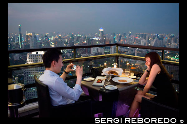 Romantic dinner. Banyan Tree Rooftop Vertigo & Moon Bar, Restaurant, , Bangkok , Thailand. View of the city, Vertigo Bar and Restaurant, roof of the Banyan Tree Hotel, at dusk Bangkok, Thailand, Asia. Reaching for the clouds at Vertigo and Moon Bar on the 61st floor of the Banyan Tree hotel is one of the best ways to end a long day in Bangkok. There is no lack of rooftop bars in town but Vertigo has always been amongst the favourites. With an unusual narrow and elongated shape, the entire top of the building is occupied by both the bar and the restaurant and gives the unusual impression of being aboard a spaceship in the sky. Located on Sathorn road, a very large busy avenue peppered with tall glass and metal skyscrapers and not far from the Lumpini park and Silom area, Banyan Tree is a name often associated with luxury. The hotel and its unusual design might be a bit aging, but once you step inside the lobby, you’re still aware of this sophisticated yet relaxed atmosphere. From the lobby, a fast lift will take you to the 60th floor where you find the popular Bai Yun Chinese restaurant, walk pass it and up a flight of stairs that leads you outdoors. You better not be afraid of heights, as Bangkok will be laid out below you the moment you step outside. What surprises people the most is to walk out on a rooftop with absolutely nothing above you. Often similar venues would have a roof or a wall or even the continuing structure blocking part of the view. Not here… the designers have successfully eliminated anything that could block your vision and the impression is accentuated by the rather slim appearance of the building… it just appears narrow of course; there’s still a 327 room hotel underneath! But the elongated shape hosting the Moon Bar on one end and the Vertigo restaurant is certainly impressive; this spaceship has an open-air bridge connecting the two areas hovering majestically above the city.