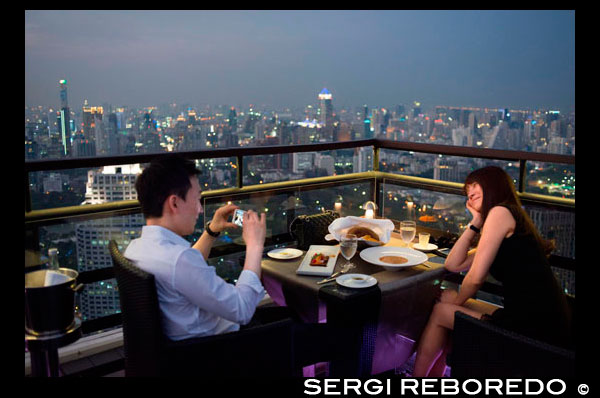 Sopar romantica. Banyan Tree terrat Vértigo i Moon Bar, Restaurant ,, Bangkok, Tailàndia. Vista de la ciutat, Vértigo Bar i Restaurant, terrat de l'Hotel Banyan Tree, al vespre a Bangkok, Tailàndia, Àsia. Arribant els núvols en Vertigen i Moon Bar a la planta 61a de l'hotel Banyan Tree és una de les millors formes d'acabar un llarg dia a Bangkok. No hi ha falta de bars del terrat a la ciutat però Vertigo ha estat sempre entre els favorits. Amb una forma estreta i allargada inusual, tota la part superior de l'edifici està ocupat pel bar i el restaurant i fa l'efecte inusual d'estar a bord d'una nau espacial en el cel. Situat a la carretera d'Sathorn, una gran concorreguda avinguda esquitxada de got alt i gratacels metàl·lics i no molt lluny del parc Lumpini i zona de Silom, Banyan Tree és un nom associat sovint amb el luxe. L'hotel i el seu disseny inusual podria ser un envelliment poc, però una vegada que entres al vestíbul, segueixes sent conscient d'aquest ambient sofisticat i relaxat. Des del vestíbul, ascensor ràpid us portarà al pis 60 on es troba el popular restaurant Bai Yun xinès, camina passar i pujar un tram d'escales que condueix a l'aire lliure. Serà millor que no tenir por a les altures, com Bangkok serà exposat a baix el moment que pas fora. El que sorprèn a la gent més és per sortir en un terrat amb absolutament res per sobre de tu. Sovint llocs similars tindrien un sostre o una paret o fins i tot el manteniment de l'estructura de bloqueig de part de la vista. No aquí ... els dissenyadors han eliminat tot el que podria bloquejar la seva visió i la impressió es veu accentuada per l'aparició més aviat prim de l'edifici amb èxit ... només sembla estret, per descomptat; encara hi ha un hotel 327 habitació sota! Però la forma allargada que allotja el Bar Lluna en un extrem i el restaurant Vertigo és sens dubte impressionant; aquesta nau espacial té un pont a l'aire lliure que connecta les dues àrees surant majestuosament sobre la ciutat.