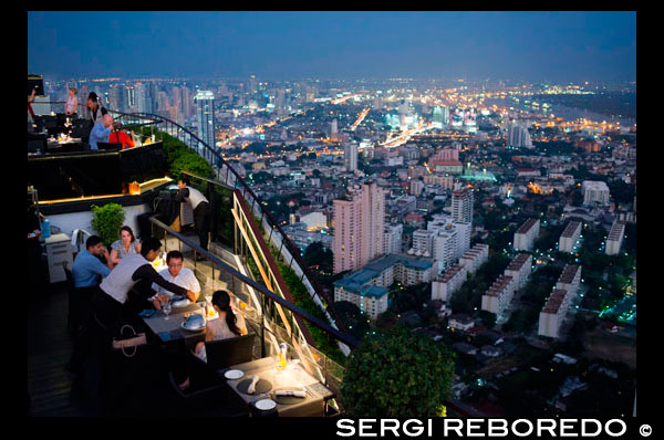 Paisaje, vistas. Banyan Tree azotea Vértigo y Moon Bar, Restaurante,, Bangkok, Tailandia. Vista de la ciudad, Vértigo Bar y Restaurante, azotea del Hotel Banyan Tree, al anochecer en Bangkok, Tailandia, Asia. Alcanzando las nubes en Vértigo y Moon Bar en la planta 61ª del hotel Banyan Tree es una de las mejores formas de terminar un largo día en Bangkok. No hay falta de bares de la azotea en la ciudad pero Vertigo ha sido siempre entre los favoritos. Con una forma estrecha y alargada inusual, toda la parte superior del edificio está ocupado por el bar y el restaurante y da la impresión inusual de estar a bordo de una nave espacial en el cielo. Situado en la carretera de Sathorn, una gran concurrida avenida salpicada de vaso alto y rascacielos metálicos y no muy lejos del parque Lumpini y zona de Silom, Banyan Tree es un nombre asociado a menudo con el lujo. El hotel y su diseño inusual podría ser un envejecimiento poco, pero una vez que entras en el vestíbulo, sigues siendo consciente de este ambiente sofisticado y relajado. Desde el vestíbulo, ascensor rápida le llevará al piso 60 donde se encuentra el popular restaurante Bai Yun chino, camina pasar y subir un tramo de escaleras que conduce al aire libre. Será mejor que no tener miedo a las alturas, como Bangkok será expuesto abajo el momento que paso fuera. Lo que sorprende a la gente más es para salir en una azotea con absolutamente nada por encima de ti. A menudo lugares similares tendrían un techo o una pared o incluso el mantenimiento de la estructura de bloqueo de parte de la vista. No aquí ... los diseñadores han eliminado todo lo que podría bloquear su visión y la impresión se ve acentuada por la aparición más bien delgado del edificio con éxito ... sólo parece estrecho, por supuesto; todavía hay un hotel 327 habitación debajo! Pero la forma alargada que aloja el Bar Luna en un extremo y el restaurante Vertigo es sin duda impresionante; esta nave espacial tiene un puente al aire libre que conecta las dos áreas flotando majestuosamente sobre la ciudad.