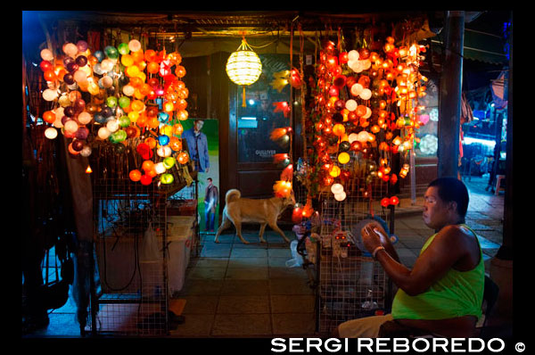 Color lamps seller in khao san road. Bangkok. Khaosan Road or Khao San Road is a short street in central Bangkok, Thailand. It is in the Banglamphu area of (Phra Nakhon district) about 1 kilometre (0.62 mi) north of the Grand Palace and Wat Phra Kaew. "Khaosan" translates as "milled rice", a reminder that in former times the street was a major Bangkok rice market. In the last 20 years, however, Khaosan Road has developed into a world famous "backpacker ghetto". It offers cheap accommodation, ranging from "mattress in a box" style hotels to reasonably priced 3-star hotels. In an essay on the backpacker culture of Khaosan Road, Susan Orlean called it "the place to disappear". It is also a base of travel: coaches leave daily for all major tourist destinations in Thailand, from Chiang Mai in the north to Ko Pha Ngan in the south, and there are many relatively inexpensive travel agents who can arrange visas and transportation to the neighbouring countries of Cambodia, Laos, Malaysia, and Vietnam. Khaosan shops sell handcrafts, paintings, clothes, local fruits, pirated CDs, DVDs, a wide range of fake IDs, used books, plus many useful backpacker items. During late evening, the streets turn into bars and music is played, food hawkers sell barbecued insects, exotic snacks for tourists, and there are also locals flogging ping pong shows. There are several pubs and bars where backpackers meet to discuss their travels. The area is internationally known as a center of dancing, partying, and just prior to the traditional Thai New Year (Songkran festival) of 13-15 April, water splashing that usually turns into a huge water fight. One Thai writer has described Khaosan as "...a short road that has the longest dream in the world".[2] A Buddhist temple under royal patronage, the centuries-old Wat Chana Songkram, is directly opposite Khaosan Road to the west, while the area to the northwest contains an Islamic community and several small mosques.