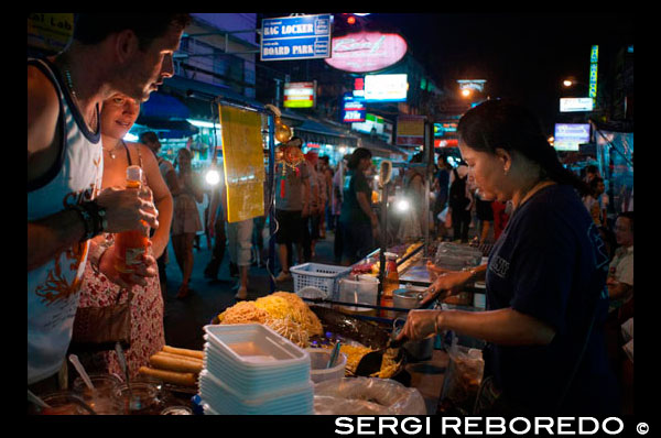 khao san road, noodles street stall. Food stall. Bangkok. Khaosan Road or Khao San Road is a short street in central Bangkok, Thailand. It is in the Banglamphu area of (Phra Nakhon district) about 1 kilometre (0.62 mi) north of the Grand Palace and Wat Phra Kaew. "Khaosan" translates as "milled rice", a reminder that in former times the street was a major Bangkok rice market. In the last 20 years, however, Khaosan Road has developed into a world famous "backpacker ghetto". It offers cheap accommodation, ranging from "mattress in a box" style hotels to reasonably priced 3-star hotels. In an essay on the backpacker culture of Khaosan Road, Susan Orlean called it "the place to disappear".  It is also a base of travel: coaches leave daily for all major tourist destinations in Thailand, from Chiang Mai in the north to Ko Pha Ngan in the south, and there are many relatively inexpensive travel agents who can arrange visas and transportation to the neighbouring countries of Cambodia, Laos, Malaysia, and Vietnam. Khaosan shops sell handcrafts, paintings, clothes, local fruits, pirated CDs, DVDs, a wide range of fake IDs, used books, plus many useful backpacker items. During late evening, the streets turn into bars and music is played, food hawkers sell barbecued insects, exotic snacks for tourists, and there are also locals flogging ping pong shows. There are several pubs and bars where backpackers meet to discuss their travels. The area is internationally known as a center of dancing, partying, and just prior to the traditional Thai New Year (Songkran festival) of 13-15 April, water splashing that usually turns into a huge water fight. One Thai writer has described Khaosan as "...a short road that has the longest dream in the world".[2] A Buddhist temple under royal patronage, the centuries-old Wat Chana Songkram, is directly opposite Khaosan Road to the west, while the area to the northwest contains an Islamic community and several small mosques.