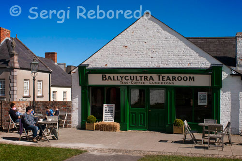 Ulster Folk & museu del transport. Ballycultra edificis de la ciutat. Ballycultra un saló de te. Tes Esmorzars de cafè. Aquestes quatre cases van ser construïdes a la dècada de 1870 com la ciutat de Dromore ampliat. El 1903, la casa més gran en l'extrem de la terrassa, amb dues habitacions en planta baixa, estava ocupada per un sabater, José Robert Magill. El seu negoci de reparació de calçat es va dur a terme a la sala i Robert, la seva dona i els seus sis fills van ocupar la cambra del fons i les habitacions de dalt. L'emprenedor Sr Magill va adquirir la casa del costat, el 1913 i va establir un negoci de reparació de bicicletes petites. Les cases eren originalment part d'una terrassa gran al carrer que va portar a Dromore Presbiteriana de casa de reunió, o l'església. Les altres cases a la terrassa són més petits, amb una sala de planta baixa, individual i una habitació de dalt i la seva distribució de la sala és una reminiscència de les cases tradicionals en el camp dels voltants. Encara que les cases eren petites, els ocupants entre 1901 i 1911 van ser totes les famílies individuals, cada un dels quatre o cinc persones. Molts dels habitants del carrer treballar a les fàbriques de roba locals. El taller de fusteria a l'extrem de la terrassa és típic de les empreses d'artesania local, que atén les necessitats d'una ciutat petita. Ubicació original: Meeting Street, Dromore, Comtat de Down