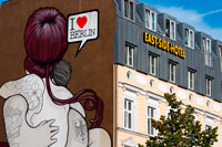 M'encanta Berlín. East Side Hotel davant l'East Side Gallery. La East Side Gallery és un tram de 1,3 quilòmetres de llarg pintada de l'antic mur de Berlín al llarg del Mühlenstraße a l'antic Berlín Est. És la major galeria a l'aire lliure del món, amb més d'un centenar de pintures murals originals. Galvanitzat pels esdeveniments extraordinaris que estaven canviant el món, artistes de tot el món van acudir a Berlín després de la caiguda del Mur, deixant un testimoni visual de l'alegria i l'esperit d'alliberament que va sorgir en aquell moment. Murals havien estat anteriorment un punt culminant per als visitants i una atracció de Berlín durant anys, però eren només per ser trobat a la banda occidental del mur. Els artistes van transformar el formigó gris rearticular això en una expressió duradora de la llibertat i la reconciliació.