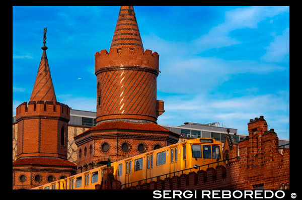 Germany, Berlin, the Oberbaumbrücke bridge that links Kreuzberg and Friedrichshain districts over the Spree river. The Oberbaum Bridge (German: Oberbaumbrücke) is a double-deck bridge crossing Berlin's River Spree, considered one of the city landmarks. It links Friedrichshain and Kreuzberg, former boroughs that were divided by the Berlin Wall, and has become an important symbol of Berlin’s unity. The lower deck of the bridge carries a roadway, which connects Oberbaum Straße to the south of the river with Warschauer Straße to the north. The upper deck of the bridge carries Berlin U-Bahn line U1, between Schlesisches Tor and Warschauer Straße stations. The bridge appears prominently in the 1998 film Run Lola Run. The bridge is built on the former boundary of the municipal area with its rural environs, where an excise wall was built in 1732. A wooden drawbridge was built as part of the wall; it served as a gate to the city. The name Oberbaumbrücke stemmed from the heavy tree trunk, covered in metal spikes, that was used as a boom to block the river at night to prevent smuggling. (Baum means tree or wooden beam in German; thus the name means something like "Upper [Upstream] Tree Bridge"; there was another tree-trunk barrier at the western end of the contemporary city limits, close to today's Unterbaumstraße (lit. in English: Lower [Downstream] Tree Street.)