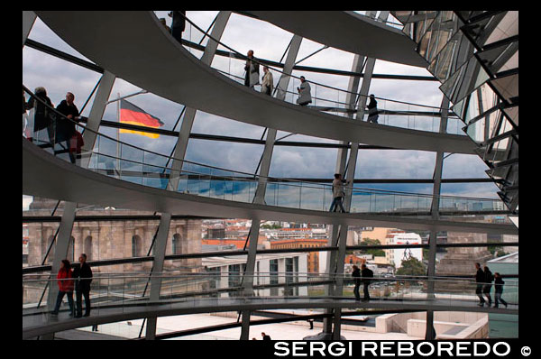 Mirrored cone inside the Reichstag Dome, in the german parlament (Reichstag), Berlin. The current Reichstag dome is an iconic glass dome constructed on top of the rebuilt Reichstag building in Berlin. It was designed by architect Norman Foster and built to symbolize the reunification of Germany. The distinctive appearance of the dome has made it a prominent landmark in Berlin. The Reichstag dome is a large glass dome with a 360 degree view of the surrounding Berlin cityscape. The debating chamber of the Bundestag, the German parliament, can be seen down below. A mirrored cone in the center of the dome directs sunlight into the building. The dome is open to the public and can be reached by climbing two steel, spiraling ramps that are reminiscent of a double-helix. The glass dome was also designed by Foster to be environmentally friendly. Energy efficient features involving the use of the daylight shining through the mirrored cone were applied, effectively decreasing the carbon emissions of the building. The futuristic design of the Reichstag dome makes it a unique landmark, and symbolizes Berlin's attempt to move away from a past of Nazism and Communism and instead towards a future with a heavier emphasis on a united, democratic Germany.