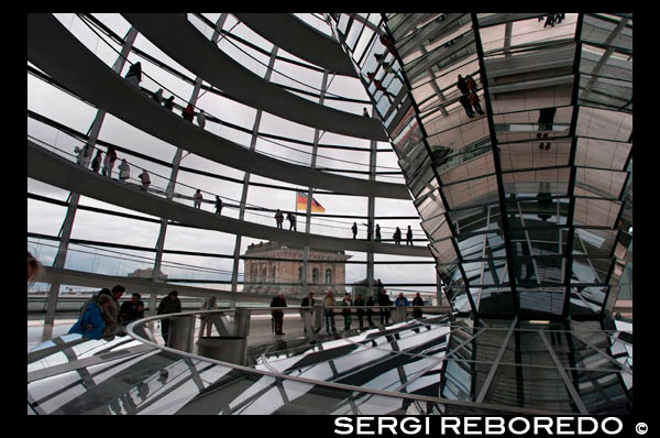 Mirrored cone inside the Reichstag Dome, in the german parlament (Reichstag), Berlin. The current Reichstag dome is an iconic glass dome constructed on top of the rebuilt Reichstag building in Berlin. It was designed by architect Norman Foster and built to symbolize the reunification of Germany. The distinctive appearance of the dome has made it a prominent landmark in Berlin. The Reichstag dome is a large glass dome with a 360 degree view of the surrounding Berlin cityscape. The debating chamber of the Bundestag, the German parliament, can be seen down below. A mirrored cone in the center of the dome directs sunlight into the building. The dome is open to the public and can be reached by climbing two steel, spiraling ramps that are reminiscent of a double-helix. The glass dome was also designed by Foster to be environmentally friendly. Energy efficient features involving the use of the daylight shining through the mirrored cone were applied, effectively decreasing the carbon emissions of the building. The futuristic design of the Reichstag dome makes it a unique landmark, and symbolizes Berlin's attempt to move away from a past of Nazism and Communism and instead towards a future with a heavier emphasis on a united, democratic Germany.