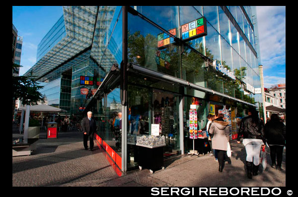 Shops behind glass facades, Neues Kranzler Eck, Berlin-Charlottenburg, Berlin, Germany, Europe. Neues Kranzler Eck Neues Kranzler Eck Linking tradition and modernity  The architecture is daring: the glass wall towers sixty metres high behind the traditional Kranzler Eck, stretching all the way to Kurfürstendamm. The Neue Kranzler Eck, designed by brilliant architect Helmut Jahn, is one of the highlights of the western city district. Surrounding the well-preserved Café Kranzler with its striking awning from the 1950s, a modern shopping centre has been developed with branches of well-known brands, small boutiques and a BERLIN ‘Infostore’.  As the famous meeting point in City West, the NEUES KRANZLER ECK, located directly on the renowned Ku’damm, invites locals and tourists to stroll, shop and relax in this exciting venue. Around 20 shops including famous fashion houses such as „KARSTADT sports“, „Buffalo Boots", „Mango“, „Schuhhof“, „H&M“, „Gerry Weber“, „Jack Wolfskin“ and „Camp David“, as well as the bureau de change,  „Exchange” with jeweller’s shop,  „Vodafone”, the  „BERLIN store”,  „BERLIN tourist information”, „AMPELMANN Shop & Café”, „Strauss Innovation”, create a real shopper’s paradise. You can purchase natural skin care, beauty & cosmetics at „Kiehl's Since 1851" and Meissen china from the MEISSEN Outlet. The „Wiener Feinbäckerei” bakery, „Super Cut“ hairstylists, „Dean and David" complete the line-up. Stop off to enjoy an art of traditional coffees in the legendary  „Café Kranzler”.