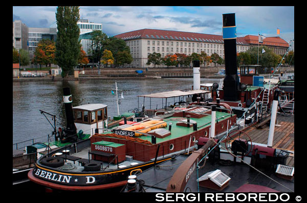 Old Harbour from Inselbruecke, Berlin, Germany. The banks of the Spree in Friedrichshain and the East Harbour offer one of the liveliest and most pleasant ways down to the river to be found in the central city area. The docks are a narrow ribbon, extending one or two kilometres between the river and the six-lane traffic route that accompanies the Spree from the Jannowitzbrücke (Jannowitz Bridge) to the Stralauer Halbinsel (peninsula).  Although there are still gatehouses, visitors can make their way, undisturbed, to the railway tracks, warehouses, loading ramps and huge travelling cranes which load the freight boats with scrap or building materials. In one of the two brick buildings used for management and administration, there was a light friendly canteen with a small summer garden going down to the Spree. This stretch of the river is more imposing and busier than elsewhere, with the Landwehr Canal on the opposite bank, Kreuzberg-Treptow Ufer, and the flood drains joining it. Before the Wall came down, the docks were an important place for handling goods for the major building sites on the edge of East Berlin. Today, activity is considerably reduced. Dandelions and camomile squeeze between the asphalt and railway tracks, and the rare bouts of industrial work merge with the atmosphere of the river to form an unbelievably romantic cityscape. The end of the idyllic industrial cityscape between Oberbaumbrücke and Elsenbrücke, (predicted for 2007), has long since come to pass. Music and fashion have taken over the old buildings and turned them into studios and showrooms, lofts and bistros. The symmetry of the old grounds, composed of two administration buildings in the centre with flat, long store-rooms to either side, has made way for new blocks and glass extensions. But for anyone mourning the loss of the old harbour canteen described above, they may find some solace in the former cold storage house, where the music giant Universal, has been based since 2002. Its canteen, which has a lovely interior design and a terrace right over the river Spree, is open to the public and is one of the nicest in the city.