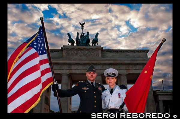 Actors vestits de soldats nord-americans i soviètics posen per fotos amb els turistes davant de la Porta de Brandenburg a Berlín , un dels monuments més emblemàtics de la Guerra Freda . La Porta de Brandenburg és la marca comercial de Berlín . L'entrada principal a la ciutat , envoltat per la paret des de fa trenta anys, era coneguda arreu del món com un símbol de la divisió de la ciutat i de la divisió del món en dos blocs de poder . Visitants internacionals d'avui dia a la Pariser Platz vénen a tornar a experimentar aquesta primera porta d'entrada a la ciutat , i gaudir de la llibertat llargament negat a caminar a través d' aquesta magnífica obra d'art i mirar de prop. Va ser construït com la més gran d'una sèrie de portes de la ciutat que constitueixen els passos a través de la paret de duanes que envolta la ciutat a finals del segle XVIII . És l'única porta que va sobreviure , ja que constitueix la terminació monumental de l'avinguda Unter den Linden , el famós bulevard de til · lers que conduïa directament a la residència dels reis de Prússia fins a la destrucció del castell de la ciutat . Tota la construcció i ornamentació de la porta reflecteixen la importància extraordinària que va ser concedit pels seus constructors . L'arquitecte seleccionat com a model per al seu disseny els Propileus d'Atenes , el hall d'entrada monumental de l'Acròpoli . Així com els Propileus va portar a un santuari del món antic , aquesta porta era representar l'accés a la ciutat més important del regne de Prússia. Aquesta referència a l'Antiguitat la va convertir en l'estructura que es va fundar l'edat clàssica de l'arquitectura a Berlín , una època que va portar a la ciutat el seu sobrenom de " Spreeathen " (" Atenes del Spree " - riu de Berlín es diu el Spree ) . L'escultor més important de Berlín durant aquest període va dur a terme el programa d'acompanyament d'explicació visual . La Porta de Brandenburg està coronat per una quadriga que representa la deessa de la victòria " , que porta la pau" , que marxa a la ciutat . El relleu al pedestal de la retrata de nou amb els seus assistents . Les personificacions de virtuts com l'amistat i l'art de governar es representen , juntament amb els símbols de les arts i les ciències , perquè fan que una ciutat com Berlín floreixen en temps de pau . Relleus amb les gestes d'Hèrcules en els passatges al · ludeixen a l'època de les guerres i el posterior període de reconstrucció , durant els quals Federico II va fer Prússia en una potència europea i va establir les bases per al floriment del comerç i l'artesania . Així doncs, la porta és també un monument per al rei que va morir uns anys abans de la seva construcció.