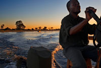 Navegando al atardecer en una lancha rápida durante el safari acuático en el campamento Eagle Island Camp de Orient Express, en las afueras de la Reserva de Animales de Moremi, en Botswana. Mbiroba es un camping que también tiene cabañas tradicionales y bungalows. Está a orillas del delta del Okavango y su mayor atractivo son los viajes en Mokoro o canoas tradicionales, que se gestionan a través de la asociación 'Okavango Poler's Trust'. Se hacen excursiones de uno o dos días y adentrarse en las profundidades del delta para disfrutar de su fauna, paisajes y calma absoluta solo rota por las llamadas de los hipopótamos al atardecer. Durante la excursión por el Delta hay que tener en cuenta que es una zona llena de animales salvajes y tener mucho cuidado de no molestarles, especialmente a los hipopotamos durante el viaje en Mokoro. El Delta del Okavango es uno de los más grandes del planeta, con 15.000 kilómetros cuadrados. A este sistema de lagos, bañados, ríos y canales llegan millones de especies en estado silvestre. Un banquete visual para los fanas de los animales.