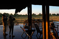 Un par de turistas en el porche exterior del campamento Khwai River Lodge de Orient Express en Botswana, en el interior de la Reserva Salvaje de Caza Moremi. Khwai River Lodge está situado a orillas del Río Khwai, junto a la Reserva de Caza Moremi y en las afueras del Delta de Okavango. Khwai River Lodge es uno de los hotel-safari más antiguos de Botswana, abierto a finales de los años 60 por el fallecido Sir Seretse Khama, primer presidente de la Botswana independiente. Está situado sobre antiguas plantas indígenas e higueras, a ocho kilómetros al noroeste de la puerta norte de la Reserva Salvaje de Caza Moremi. Además, domina con sus vistas el río Khwai y es el lugar perfecto para observar el paisaje perenne de la sabana africana y descubrir sus variadas especies de aves. El río Khwai forma parte del Delta de Okavango. El campamento se encuentra en  Moremi, una de las reservas de mayor majestuosidad de África que presume de albergar los ejemplares más grandes, rápidos, relucientes y bellos, como los abundantes cocodrilos e hipopótamos que moran en sus tierras o las manadas de cebras y antílopes que rasgan satisfechas la superficie de la hierba. Allí donde éstos se alimentan, acechan todo tipo de depredadores: leones, leopardos y panteras, hienas y perros salvajes. 