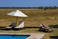 Una turista toma el sol en bañador junto a la piscina en el campamento Khwai River Lodge de Orient Express en Botswana, en el interior de la Reserva Salvaje de Caza Moremi. Khwai River Lodge está situado a orillas del Río Khwai, junto a la Reserva de Caza Moremi y en las afueras del Delta de Okavango. En su ribera y humedales habitan numerosas especies -hipopótamos, elefantes, leones, leopardos, antílopes y abundantes clases de pájaros- a sólo unos metros de los campamentos, abiertos lateralmente por el comedor y con vistas desde el césped de la piscina.  El campamento dispone de 15 magníficas tiendas de lujo, una suite con bañera de cobre exterior de estilo Victoriano, ducha, piscina propia y una terraza aislada.  Cada tienda tiene un espacio sombreado con una hamaca y cómodas sillas con vistas al Río Khwai y su abundante vida animal. Equipado con todo tipo de comodidades modernas -incluido mini-bar y sistema de ventilación de aire acondicionado- las tiendas fueron decoradas  por el famoso diseñador Graham Viney. 