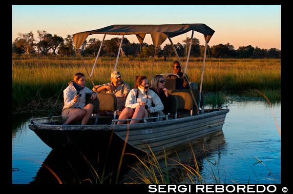 Los turistas disfrutas de los últimos rayos de sol en el safari acuático efectuado desde el campamento Eagle Island Camp de Orient Express, en las afueras de la Reserva de Animales de Moremi, en Botswana. El río Okavango nace en Angola y después de recorrer casi 1000 kms se divide en infinidad de riachuelos, canales y lagunas formando el área llamada Delta del Okavango (ya en Botswana). 15.000 kilómetros cuadrados ocupa este increible regalo de la naturaleza llena de aves de todos los colores, hipopótamos, cocodrilos, impalas, cebras, elefantes, búfalos, jabalíes... así como nenúfares, papiros y de la más variada fauna.  Los safaris se hacen tanto a pie como en lancha.  También se visitan aldeas dónde contemplar la vida humilde de sus habitantes : casas de madera compartiendo un surtidor de agua y sin luz. Además del moroko dónde llevar a los turistas, sus fuentes de ingreso son pequeños huertos, la pesca  y trabajos de artesanía.  RECOMENDACIONES Si lo que te interesa ver son grandes mamíferos, la mejor época para visitar el delta es de mayo a octubre, cuando las aguas están en niveles bajos. Si prefieres una vegetación verde y rabiosa cubierta de aves, la mejor época es la de las lluvias, de noviembre a abril. Los alojamientos situados en el corazón del delta son privados y son muy caros. La alternativa son los campamentos y los lodges del gobierno que se encuentran en la península de Moremi.  CURIOSIDADES Se dice que los leones del Delta del Okavango son los únicos nadadores, que en época de crecidas del agua se ven obligados a hacerlo si se quedan aislados en islotes para cazar a sus presas más comunes: los antílopes y los impalas. Los mokoros son una especie de canoas utilizados por los habitantes de la zona para desplazarse por los canales. Construida con madera se mueven lentamente impulsadas por la fuerza del hombre.