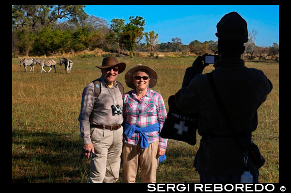 Una pareja de turistas se fotografía con las cebras de fondo en el safari a pie realizado en las inmediaciones del campamento Eagle Island Camp de Orient Express, en las afueras de la Reserva de Animales de Moremi, en Botswana. Tras otear el horizonte, Botsualo, nuestro guía, nos autoriza a bajar para instalar las tiendas de campaña e iniciar una caminata al atardecer. “No tenéis que preocuparos, en esta zona del delta no hay depredadores” nos dice cuando ve nuestras caras, entre asustadas e ilusionadas por caminar por esa zona. Normalmente en los parques naturales no se puede caminar pero en zonas determinadas, con guía sí. Esta es una. Sigilosos avanzamos reconociendo enormes pisadas de elefantes y excrementos de monos, hasta una zona de pastos donde varios grupos de cebras, ñúes y antílopes pastan a sus anchas. Descubrimos un esqueleto de jirafa. Más lejos, junto a un enorme baobab, un elefante solitario. No podemos acercarnos demasiado pero el estar allí, sin la protección del coche, en plena naturaleza, en su terreno, es una sensación maravillosa y liberadora a partes iguales. A medida que cae el sol nos retiramos. Aunque sea la mejor hora para observar los animales tenemos que regresar al campamento, a encender el fuego y cocinar la cena. No hemos hecho ningún esfuerzo, pero la naturaleza en estado puro parece que hace entrar el hambre. Tras la cena, la humedad de la noche trae el frío a nuestros huesos aunque por suerte tenemos las ascuas para calentarnos. En el silencio casi total cualquier ruido es perceptible, así que nos quedamos mudos al oír, a apenas unos metros de distancia, ruido de agua, pisadas en barro, ramas rompiendo, árboles zarandeados… “No os mováis ni encendáis ninguna luz”, nos dijo Botsualo dejándonos solos mientras salía a mirar, sigiloso. Dedujimos que un ruido así solo podía ser un elefante. A medida que se alejaba el ruido el guía regresó con un enorme palo en la mano. “Los elefantes son los animales más peligrosos. Solo duermen 3 ó 4 horas y siguen paseándose por la noche. Y pueden no ver tu tienda de campaña y…” nos dijo, dejando la frase inacabada. “Una vez”, prosiguió, “para espantar a uno tuve que golpearlo porque se había metido, muy molesto, en un campamento, donde teníamos antorchas”. Así es, en el delta, durmiendo en algún islote, no hay vallas ni protecciones. Es pura naturaleza, su territorio, esa es la belleza. Y algo así no tiene precio.