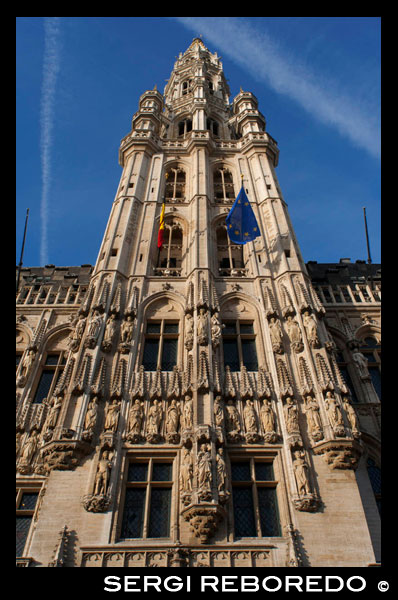 Hôtel de Ville. L'edifici de l'ajuntament, que ocupa la façana sud-oest, és l'únic edifici medieval que queda avui en dia en peu i va ser construït entre 1402 i 1455 probablement per Jean Bornoy. La seva torre, asimètrica pel que fa a l'edifici, de 96 metres d'altura, és d'estil gòtic i va ser dissenyada per Jean van Ruysbroeck en 1449. Una llegenda urbana compte que l'arquitecte, en comprovar el suposat error que la torre no quedés situada al centre, va pujar a aquesta i es va llançar al buit, encara que en veritat ambdues parts no van ser construïdes a la vegada. A sobre de la torre es troba una alta estàtua de bronze de 5 metres de l'arcàngel Miquel, patró de Brussel · les, matant a un drac o dimoni. La part esquerra de l'ajuntament consta de dotze arcs, inclòs el campanar, que representen l'alquímia humida en dotze etapes. En canvi, el costat dret té únicament amb set arcs, comptant el campanar, que simbolitzen la nova alquímia seca, en set etapes. La façana està decorada amb nombroses estàtues que representen els nobles, els sants i figures al · legòriques. Contrapicat L'AJUNTAMENT DE BRUSSEL · GRAND PLACE