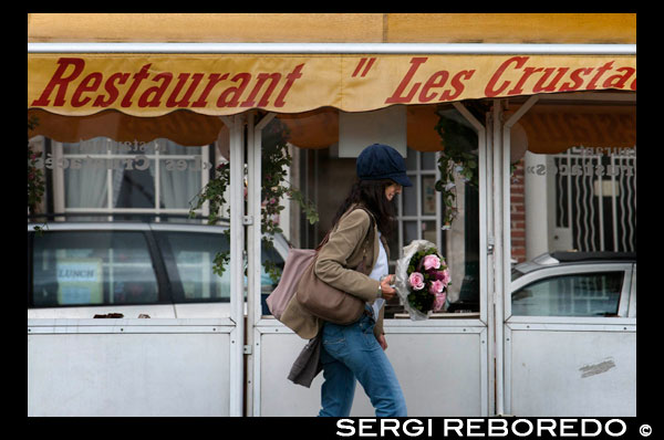 Una chica con flores pasea junto a la zona de restaurantes de Ste-Cathérine y St Géry. La zona de Ste- Cathérine resulta muy animada, sobretodo por la noche. Gran cantidad de marisquerías dispuestas una detrás de otra se disputan a los clientes, en un barrio popular y marcadamente flamenco. Sus terrazas y salones para degustar el chocolate ralentizan el ritmo de una ciudad vibrante. Saint-Géry, situado más al sur, toma su nombre de una antigua parroquia, en este caso, la de san Gaugerico de Cambrai (ca. 550-ca. 626), que fue obispo de esa ciudad de las Flandes francesas. Antes de la importante transformación urbanística, Saint-Géry era una isla formada por el río Senne. La isla formaba un gran círculo que iba desde la antigua iglesia de las Clarisas hasta la actual bolsa, junto a otra segunda isla más pequeña cercana a la plaza Fontainas, donde el río bordeaba la iglesia de Nuestra Señora del Socorro. CHICA ROMANTICA CON FLORES JUNTO AL RESTAURANTE.