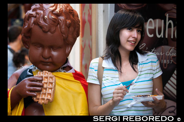 Una de les grans experiències que has de viure en visitar Bèlgica és provar les seves mundialment famosos gofres o wafles. Els trobaràs en llocs de carrer, bars, restaurants i en cada casa. Tantes varietats com ciutats belgues hagi, però tots amb una fama justificada. El gofre belga és més alt, esponjós, lleuger i saborós que el tradicional o el conegut als Estats Units. De fet la versió americana neix a partir dels belgues que van emigrar i van portar la seva típica recepta, però adaptada a les farines i paladars de l'altre costat de l'Atlàntic. Els wafles belgues són cruixents i daurats per fora, i suaus, esponjosos i blancs per dins. En realitat el conegut a la resta del món com "wafle belga" és en realitat el "wafle de Brussel · les" que va ser mal batejat durant la Fira Mundial de Nova York de 1964 per sintetitzar, i així va quedar. Com altres països europeus, la tradició belga dels gofres es remunta a l'edat mitjana quan es preparaven els "Wafers", uns pans fets amb una massa semi líquida i cuits entre unes planxes calentes sobre el foc. Del mateix origen poden haver nascut els pancakes i les crêpes. El wafle de Brussel · les ("Gaufre de Bruxelles" en francès) es caracteritza perquè la seva massa porta rovells, ous i mantega fosa, de manera que el resultat és més esponsojo que altres receptes belgues. A més, les "wafleras" de Brussel · les són més grans i profundes de manera que aquests wafles poden ocupar el plat sencer amb una més abundant guarnició de melmelades, fruites, gelat, fruits secs i / o xocolata. És les postres tradicional a Brussel · les i no t'ho pots perdre (deixa la dieta per quan tornis a casa). Un altre dels famosos wafles belgues és el de Lieja ("Gaufre de Liège" en francès). És de massa més densa i consistent que l'anterior, una mica més petit i caramel · litzat, sol acompanyar amb sucre glace. És molt comú trobar-los en els llocs de menjar de carrer perquè són més fàcils per menjar mentre camines. Els podràs veure també a la calor dels expositors als forns encara no duren molt allà perquè són un excel · lent company de les visites a peu per les ciutats quan fa una mica de fred, encara que poden menjar-se també temperats o freds. Et costaran aproximadament 3 euros, depenent del lloc i els toppings. UNA TURISTA ES FOTOGRAFIA menjant-se un gofre.