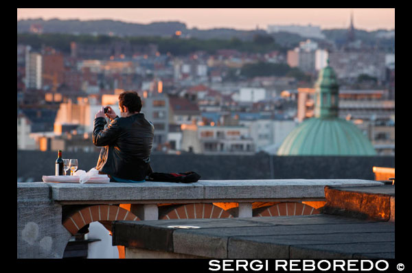 Un hombre fotografía los tejados de Marolles desde las proximidades del Palacio de Justicia. Ubicada en el centro de Marolles, barrio popular de Bruselas por excelencia, se halla la Place du Jeu de Balle o Vossenplein. Plaza también conocida con el nombre de «Mercado Antiguo». La plaza Jeu de Balle se creó en 1854, al mismo tiempo que la Rue Blaes. Entonces, se llamaba «Renard» porque lindaba con la Rue du Renard, que ya existía en el s. XV. Goza de muy buen ambiente y es popular por su rastro, que podrá visitar todas las mañanas. Se encontrará con los amantes de las antigüedades, paseantes, visitantes por un día... Sobre todo, le recomendamos que se siente en una de sus terrazas para saborear una buena cerveza y disfrutar de la música en vivo, en particular en verano. UN ROMANTICO CON VINO EN LOS TEJADOS DE MAROLLES.