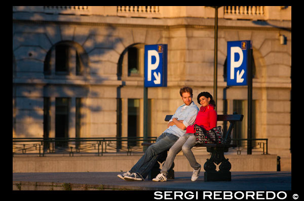 Una pareja romántica sentada en un banco en la plaza Poelaert junto al ascensor de Marolles. Para bajar desde el barrio Sablon hasta Marolles se puede utilizar un medio de transporte insólito, un ascensor gratuito íntegramente de cristal desde el que se disfruta unas espléndidas vistas sobre Marolles. Lo pusieron para salvar el desnivel que separa Les Marolles del Palacio. En la parte de arriba (plaza Poelaert) hay un mirador desde el que se ven algunos de los edificios más emblemáticos de la ciudad. LA BRUSELAS MAS ROMANTICA EN PAREJA