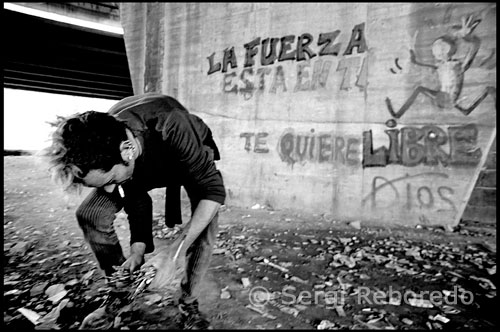 CAN TUNIS BARCELONA DRUGS. Un usuario bajo el puente de Can Tunis busca el máximo de jeringuillas usadas para poder intercambiarlas por nuevas y venderlas para ganarse su dosis. En la pared: La fuerza esta en ti. Dios te quiere Libre. Está el que acaba de ser padre, que tiene a la madre de su hijo, también drogodependiente, en el hospital tras el parto. Al bebé han tenido que suministrarle metadona, porque tiene el síndrome de abstinencia recién nacido. Él continua yendo a consumir. Y este otro caballero, que va buscando de aquí y allá hasta que reúne el necesario para una dosis. En ocasiones sirve de “conejillo de indias”, prueba el cargamento, antes de que lo adquieran los “camellos”, para constatar la calidad de la mercancía en su propia cuerpo. “Ayer les dije, que esto era una mierda, que no valía nada”. Ahora está delante mío preparándose, antes de inyectarse, sale una gota por la punta de la aguja, la contempla pasmado como quien esta viendo, la séptima maravilla, del mundo, o la Capilla Sixtima y exclama: “¡Que belleza!”, y acto seguido le pasa la lengua, y se relame complacido. No es dulce, y por tanto no ha sido cortada con Cola-cao, como pasa a menudo.   Mientras se pincha me explica, “Yo lucho porque sea legal, o que en cualquier caso pase como en Austria, o en Alemania, donde recibes asistencia inmediata, y no has de hacer colas interminables para recibir tratamiento. Quiero que mis hijos puedan decir: “Mi padre fue un yonqui, pero hizo lo que pudo por mejorar la situación...”. 