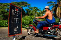 Isla Atiu. Isla Cook. Polinesia. El sur del Océano Pacífico. Un habitante de Atiu conducir con su motocicleta en las carreteras de la isla junto a un cartel de mantenerse a la izquierda. Aunque Atiu es ideal para caminar, un scooter (moto) o en bicicleta se le permiten explorar más de la isla a su propio ritmo. Atiu Villas alquila scooters, bicicletas y un jeep. Sus propios clientes tienen prioridad, pero también va a alquilar a otros. Hay otros negocios en la isla alquilar scooters, aunque también y tus acomodador pueden ayudar a organizar esto. Usted debe hacerles saber que usted puede ser que desee alquilar una moto cuando la reserva de su alojamiento. Atiu Guesthouse and Atiu Homestay también tienen un vehículo de tracción en las cuatro ruedas se alquilan. Una vez más, tendría que pagar para hacer consultas y reservar con antelación para asegurarse de que esté disponible. Si usted está haciendo algunas excursiones (muy recomendado) y algunos a pie, puede que sólo desee alquilar una moto o jeep por un día - dependiendo por supuesto de cuánto tiempo te vas a quedar en Atiu. Debido a la naturaleza pequeña y personalizada del turismo en Atiu, todos los acomodador le recogerá en el aeropuerto y le llevará a su alojamiento. Transporte puede ser en la parte posterior de un ute - viajar como un local y disfrutar de la vista! De gasolina en Atiu es caro y por lo general hay un costo para traslados al aeropuerto, aunque puede ser incluido en su paquete de alojamiento. Verifique de antemano para evitar cualquier malentendido, cuando llega el momento de pagar la cuenta. No hay transporte público o el servicio de taxi en Atiu.
