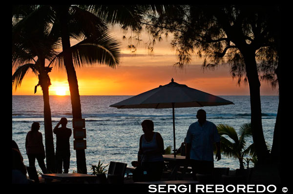 Isla de Rarotonga. Isla Cook. Polinesia. El sur del Océano Pacífico. Puesta de sol en la playa en el Hotel Crown Beach Resort & Spa. Iluminada. Los turistas tomando fotos. Sombrillas. El Crown Beach Resort se compone de 22 villas situado en acres cuatro y medio de jardines tropicales, que da a una hermosa franja de playa de arena blanca. La impresionante piscina tiene unas vistas fantásticas de la playa y el árbol de la llama serena y jardines; un lugar ideal para relajarse con un buen libro. Después de un duro día tomando el sol o bucear usted puede hacer su camino hacia el spa para un masaje terapéutico o tratamiento. Junto con desayuno tropical y té de cortesía por la tarde, este complejo cuenta con dos restaurantes: El Windjammer, que sirve deliciosos pescados del día y pan hecho en casa, y la playa lado Cabana Bar and Grill, para una cena más informal. Cada villa es cómoda autónomo que garantiza el máximo confort, lo que permite disfrutar la calidez y el encanto natural de las Islas Cook en este complejo atento.