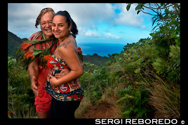 Rarotonga Island. Cook Island. Polynesia. South Pacific Ocean. A tourist with maorí tattoo takes pictures with Mr. Pa.  Ten minutes into my ascent of Rarotonga’s 413m-high ‘Needle’, a dip into the warm waters of the island’s halo-like lagoon is effortlessly topping my ‘must do’ list. But first I need to negotiate the steep and slippery tangle of roots and mini-ravines making up the first half of Rarotonga’s Cross-Island Track. Close behind me is local hiking legend Pa, carrying plenty of fresh papaya and bananas for on-the-go sustenance. Pa is a well-worn nickname, apparently borne out of the frustra- tion his German ex-wife had in pronouncing his much longer traditional name. And like his adopted moniker, everything about Pa is stripped back and simple. No shoes and no fancy hiking gear – just an innate understanding of the mountainous tropical landscape we’re traversing. His unlined face and muscular physique belie the fact that he’s more than 70 years old, testimony to his more than 3700 ascents of the peak, known more formally as Te Rua Manga. After 40 minutes uphill we reach the base of the Needle. Across a series of rugged val- leys, Pa indicates the 653m-high Te Manga, shrouded today in amorphous cloud. Pa has climbed Rarotonga’s highest mountain only a mere 800 times – compared to the Needle, it’s probably a hike he’s still getting his head around. My own headspace is filled with the green-shrouded spectacle of one of Polynesia’s most beautiful islands. Downhill from our exposed bluff, the Cross-Island Track continues for another hour through cloistered and mossy river valleys to end in the cooling haven of the Wigmore Falls.