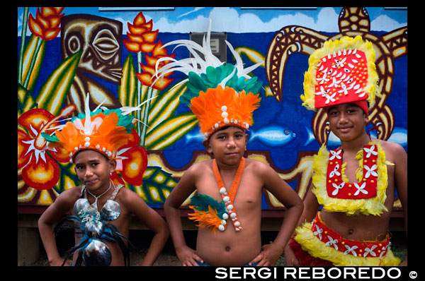 Illa de Rarotonga. Illa Cook. Polinèsia. Un grup de nens vestits com a ballarins de l'illa de cuiner al voltant dels Mercats Punanga Nui. Ballant a Illes Cook és gairebé similar a la dansa tahitiana. La diferència entre les Illes Cook i el ball tahitià és al balanceig del maluc i els peus. Dones ballarins moviment del maluc ha de ser predominantment de costat a costat i que ha d'estar desprevingut mentre fa això. Dones tahitianes ballarins moviment del maluc és majoritàriament ronda i ronda (com una rentadora) amb el taló aixecat. És una veritable prova per a romandre desprevingut mentre balancejava seus malucs. La manera d'explicar una bona ballarina és si les seves espatlles es mantenen estables, mentre ella es balancejava seus malucs. Els materials bàsics utilitzats per fer les faldilles de ball són: fulles sempre verdes (Rauti o coco frondes), tires d'escorça seca de l'arbre de hibisc salvatge (kiriau), colpejaven l'escorça (tapa) o un drap de cotó. A la cintura normalment hi ha una titi que està adornada amb tot tipus de petxines, llavors, fulles o flors. A part de ser un element decoratiu, sinó que també afegeix una mica de pes que ajuda a que el tremolor / procés tirant. Molta feina entra al vestidor que produeixen, sinó que va durar molts anys. Darrere dels ballarins són les bateries, guitarra / jugadors ukelele i cantants. Hi ha diversos instruments de percussió utilitzats. El paté (partay pronunciat) és una peça de fusta que ha estat buidat que conté una ranura a la part superior. Observeu aquests es veuen afectats, ja sigui amb un pal o dos pals en funció del tipus de rythmn vostè està tractant de produir. Les diferents longituds, tipus de fusta i el diàmetre de la paté també afecten al so. Després hi ha els tambors que són similars als bidons metàl · lics normals que es poden comprar excepte aquests són de fusta i cuir de cabra. L'enorme tambor inclinat sobre un costat produeix el so boom boom boom que se sent. Una gran part de peu tambor prim produeix un so diferent en posició vertical i es colpeja amb dos pals. Els pals que copegen es duen a terme de manera diferent, més semblant a la celebració d'un con de gelat, però amb el polze pressionat contra el con. De tota manera, és molt difícil de descriure, és millor si ho vas veure. Crec que una vegada que escolti els cops de tambor, haurà de aixecar-se i unir-se a l'espectacle també. Ells diuen que el ball de les Illes Cook és molt sensual, sigui vostè el jutge. Hi ha una sèrie de grups de dansa que es realitza en diversos hotels de l'illa. Vostè només pot veure l'espectacle, però normalment hi ha una consumició mínima d'uns NZ $ 5 (EUA $ 2.50). Assegureu-vos de seure a la davantera.