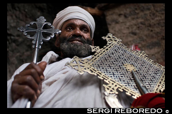 El sacerdote de la iglesia de Nakuto Lab, a las afueras de Lalibela muestra su cruz. Se desconoce el motivo por el cual fueron excavadas o construidas en el suelo, construcción que elevada la dificultad de la edificación y aumentaba los costes. Se barajan varias teorías, una de las más aceptadas es que fueron levantadas durante el auge de las peregrinaciones cristianas, siendo Jerusalén el eje o centro principal de las mismas; durante el camino, el peregrino se enfrentaba a diversos peligros y las iglesias estaban expuestas a actos de vandalismo y saqueos, tal vez este fuera el motivo por el cual, el rey Lalibela decidiera soterrar las iglesias, protegiéndolas de posibles saqueos. Poco se sabe también del proceso de construcción, no se han encontrado planos, no se conoce el nombre de los arquitectos, la duración de los trabajos, la metodología de trabajo empleada, el uso de andamios o rampas…, lo que si se sabe es la dureza del trabajo que llevó la construcción de las iglesias, donde los “obreros” realizaron un trabajo como si de la excavación de una mina se tratara. El conjunto arquitectónico de Lalibela lo conforman once iglesias excavadas en el suelo de piedra volcánica de color rojizo. Primero se labró un foso alrededor de la masa pétrea o del espacio que iba a ocupar la futura iglesia, este foso permitiría el trabajo desde el exterior a los constructores y, hoy da acceso a los templos mediante escaleras o rampas. Su simbolismo es la separación del espacio sagrado del profano. 