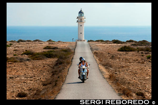 Two young motorcyclists on a long road to Es Cap de Barbaria lighthouse, in Formentera, Balears Islands. Spain. Barbaria cape formentera lighthouse road.  Es Cap de Barbaria is a heavenly place formed by rocks, bringing together a feeling of enormous loneliness and freedom. You need to visit and walk here to realise how majestic it is. Finally, if you reach the very end of it, you will see where the lighthouse stands, a place where a unique sunset can be enjoyed as the sea and sky blend in reddish tones on the horizon. If you still feel like walking, you can visit the nearby watchtower lighthouse that once protected the island from invaders.