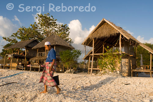 En el noroeste de la isla se encuentra el Café Diana, el mejor lugar donde ver las magnificas puestas de sol mientras se saborea un cóctel o un sabroso pancake de coco. Gili Meno. Un camino de piedra blanca, bordeado de antorchas de palma, conecta los bungalows y la zona de chill-out de Tao Kombo. Banderolas triangulares se mecen sobre largas cañas de bambú. Máscaras melanesias y la figura de un hombre de madera, arrodillado agarrándose un brazo con el otro, adornan el camino. Ese hombre parece explorar con detalle una maceta con un pequeño tronco del que salen hojas verde y una flor roja que culmina un pequeño bonsái tropical. Mira hacia el bar y la recepción, una casa de adobe guarnecida con un colchón de hoja de palma seca. Un ánfora negra con dibujos en blanco, un farol de gas y una gran maceta de la que brotan flores rosas. Todo es silencio y pájaros a media tarde, hora de la siesta para los huéspedes.