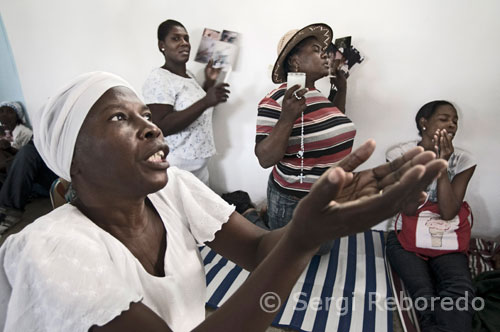 Varias mujeres rezan en el interior de la iglesia de Nuestra Señora del Monte Carmel. En las proximidades de esta zona miles de peregrinos acampan en tiendas de campaña, al raso o como huéspedes en las casas vecinas. Llegan después de muchas horas de camino a pie, en burros y en "tap tap", los coloradísimos y típicos autobuses, en busca de buena suerte y beneficios.  En el país más pobre de América, el costo de este viaje puede significar días o meses de trabajo, sacrificio que será recompensado por las bendiciones que les llegarán a los peregrinos al bañarse en las aguas místicas de la cascada de Saut d'Eau (salto de agua, en francés), en Ville Bonheur, departamento del Centro. 