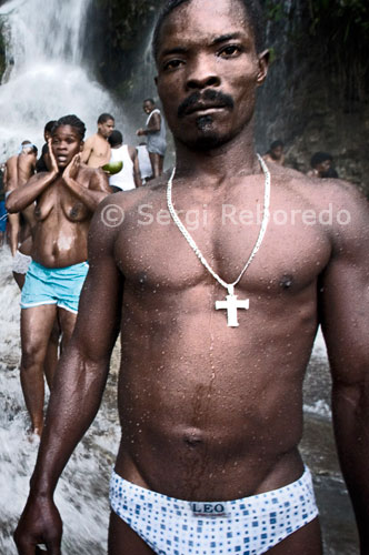 Un dels homes assistents al festival vudú de Saut d'eau que se celebra cada mes de juliol a Ville-Bonheur. El festival de vudú de Saut d'Eau se celebra cada any el 16 de juliol coincidint amb el dia en què el 1847 va aparèixer una visió de la Verge Maria. Milers de devots arribats de tots els llocs de Haiti es banyen en aquestes aigües per purificar o compra perdre al Gran Mestre que converteixi en realitat els seus somnis. El vudú és la religió majoritària de Haiti. Els adeptes necessiten ser posseïts per un esperit Iwa per poder comunicar-se amb el Gran Met, ja que aquest es encuetra molt allunyat del pla físic. A la fotografia un dels assistents muesta una creu cristiana. Els adeptes a aquest tipus de religió vudista també es consideren catòlics. El vudú està present d'una manera o altra en gairebé tot el món en Haiti. Per poder contactar amb el Gran Met es necessita un Iwa. El primer grup de Iwa són els Rada, i el seu principal color cerimonial és el blanc. En vudú també se l'associa a la màgia. Baron Samedi és l'últim recurs contra la màgia perquè, fins i tot si un malefici col.loca a una persona a la vora de la mort, pot negar-se a "cavar la fossa", de manera que aquesta persona no pot morir.