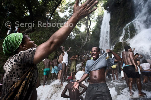 A prop de la cascada, homes i dones venen espelmes, cordons blaus i blancs que representen els lloances i imatges de la Verge dels Miracles. Els tambors toquen ritmes sincopats, i algunes persones entren en trànsit, parlant en veu alta i movent-se frenèticament. En aquest moment el Ron Barbancourt, el més típic de Haiti, entra en acció perquè la festa sigui completa. El festival de vudú de Saut d'Eau se celebra cada any el 16 de juliol coincidint amb el dia en què el 1847 va aparèixer una visió de la Verge Maria. Milers de devots arribats de tots els llocs de Haiti es banyen en aquestes aigües per purificar o compra perdre al Gran Mestre que converteixi en realitat els seus somnis. El vudú és la religió majoritària de Haiti. Els adeptes necessiten ser posseïts per un esperit Iwa per poder comunicar-se amb el Gran Met, ja que aquest es encuetra molt allunyat del pla físic. A la fotografia diverses persones resen mirant cap a la cascada mentre altres celebren aquest dia màgic. El vudú o Vodun és una religió originada a l'àrea cultural de l'Àfrica Occidental en temps prehistòrics. Es tracta d'una variant teista d'un sistema animista de creences, proveït d'un fort component màgic. Per la seva vinculació directa amb la cosmologia i els sistemes de creences neolítiques, el seu estudi resulta de gran interès en el camp de la Paleoantropologia. El vudú es compta entre les religions més antigues del món, a cavall entre el politeisme i el monoteisme. El tràfic d'esclaus cap a Amèrica va produir un fort fenomen de sincretisme entre aquesta religió arcaica i les creences cristianes dels esclavistes, així com amb les religions natives dels llocs on es va transportar als esclaus. D'aquí sorgiria el vudú haitià i un gran nombre de derivatius: la Regla de OCHA o Santeria a Cuba, el Candomblé, la Umbanda i Kimbanda al Brasil, etc. Alguns d'aquests derivats han arribat a Europa en dècades recents, sobretot de la mà d'emigrants retornats.