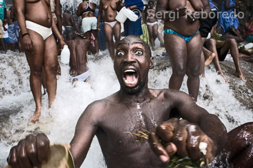 Un hombre en trance en una de las pozas de Saut d’eau. El festival de vudú de Saut d'Eau se celebra cada año el 16 de Julio coincidiendo con el día en el que en 1847 apareció una visión de la Virgen Maria. Miles de devotos llegados de todos los lugares de Haiti se bañan en estas aguas para purificarse o pra perdirle al Gran Maestro que convierta en realidad sus sueños. El vudú es la religión mayoritaria de Haiti. Los adeptos necesitan ser poseidos por un espiritu Iwa para poder comunicarse con el Gran Met, ya que este se encuetra muy alejado del plano físico. En la fotografía un adepto se encuentra poseido por el gran Iwa. Grita y sus movimientos son convulsos. El trance, momento en el que los espíritus Iwa se introducen en el cuerpo de los adeptos. Desde primeras horas , los fieles haitianos comienzan a acudir a los campos santos cargados de café, «klerec» (bebida autóctona de fuerte graduación alcohólica) y todo tipo de alimentos para ofrecer a los espíritus. El vudú, aunque no de una forma siempre visible, está presente en todos los ámbitos de la vida del país y hasta los colores azul y rojo de la bandera nacional representan a Ogou, espíritu de la guerra, el fuego y el cosmos. Previamente el rito exige que estas mujeres consagradas a la religión vudú purifiquen sus cuerpos y limpien sus órganos sexuales con el líquido que extraen de una botella repleta de hierbas antes de recibir al espíritu. Violentos movimientos, bailes y una continúa ingesta de alcohol caracterizan los momentos en los que las sacerdotisas del vudú prestan sus cuerpos a los espíritus de los muertos. La multitud, que en todo momento permanece alrededor de los sacerdotes, anima con cantos y gritos a éstos en sus contactos con el otro mundo. Otra de las características esenciales de esta religión de origen africano es su profunda fluidez, en parte porque no tiene liturgia ni teología escrita y en parte por las diversas influencias que ha tenido de otras religiones.