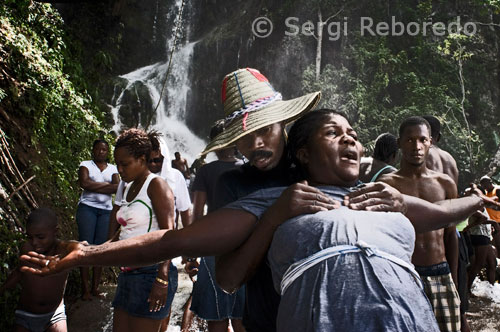 Una parella de haitians al costat de la cascada de Saut d'eau. Els oficiants voduistas haitians mostren simplicitat al vestidor i els atributs que s'insereixen en ell com clar referent religiós. No passa així entre els seus descendents que tenen jerarquies semblants, en els que s'observa, per contra, una sobrecàrrega en els elements visibles. Així, mostren múltiples tipus de collarets amb comptes de llavors amb les que es fan els que es venen en alguns establiments públics. Entre elles s'insereixen altres objectes, com xapes de clauers i les anomenades "llàgrimes" de vidre de les làmpades d'aranya. En un collaret observem desenes de carretel de fils de cosir de diferents colors. És evident que a la funció cultural s'afegeixen elements decoratius. No falten en aquests atributs medalles d'or amb imatges de santa Bàrbara o de la verge de la Caritat del Coure.