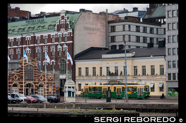 Hèlsinki. Vistes genrales de la ciutat amb el tramvia i el mercat de fons al carrer Eteläranta. Visita al mercat. Al costat del mercat exterior del port, trobaràs el mercat cobert. És un bon lloc per provar el peix local o per comprar embotits de ren. Plaça del Mercat Vell Aquesta zona de la ciutat de Hèlsinki, és potser la més bulliciosa i aglomerada de la ciutat. Les raons són evidents, d'una banda és el nus del qual parteixen els carrers comercials més importants, també és punt de partida dels feries que porten a Suomennlina i els vaixells que realitzen passejos per la costa. A part l'envolten monuments com la Catedral, l'Església Uspenski, el Palau Presidencial i la Cort Suprema, i és, en si centre del petit comerç i dels llocs de menjar, artesanies i fruites de temporada que es realitzen a l'estiu, que es converteixen en artesania i rebosteria nadalenca al desembre. Després de pensar que no hi havia gairebé ningú a la ciutat, arribar a la plaça i veure plena de gent i de turistes (en aquesta plaça es troba la principal oficina d'informació de Hèlsinki) va ser una mica xocant, ja que havia estat gaudint de la tranquil · litat de la ciutat des de primeres hores del matí. Però ja se sap, que els molls i ports, d'aquestes ciutats eminentment marítimes són així, el focus de la vida diària, sobretot a l'estiu. Ah, una dada important. L'obelisc que veiem va ser el primer monument públic que es va erigir a Hèlsinki el 1835. La seva part superior presenta l'àguila imerial russa, record de la visita del Tsar Nicolas i la seva dona dos anys abans.