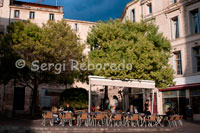 Ambiente en los muchos cafes cercanos a la iglesia de Sainte Anne. Rue Ste Anna.  En este pintoresco barrio, entre el conservatorio de música y la iglesia de Santa Ana, se pueden ver algunos talleres de luthiers, los artesanos que se dedican a la construcción y reparación de instrumentos de cuerda.Continuamos por las animadas calles peatonales que ahora ya están a reventar y bajando por la rue de la Loge llegamos a la Plaza de la Comedia, centro vital y punto de encuentro de los habitantes de la ciudad. Este gran espacio está presidido por la Ópera Comedia y rodeado de otros edificios de solera. En el centro de la plaza se encuentra una copia de la fuente de las tres gracias – Aglaya, Eufrósine y Talía – uno de los emblemas de Montpellier. La estatua original, creada por Etienne Antoine en 1970, se encuentra en el hall de la Ópera Comedia.