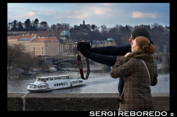 Praga romántica. Un pareja de fotografía en el Puente de Carlos.  El Puente de Carlos (en checo Karl?v most) es el puente más viejo de Praga, y atraviesa el río Moldava de la Ciudad Vieja a la Ciudad Pequeña. Es el segundo puente más antiguo existente en la República Checa. La necesidad de un nuevo puente surgió luego de que el viejo Puente de Judith fuera destruido por una inundación en 1342. Este puente de estilo románico había sido bautizado en honor a la esposa del rey Ladislao I. Astrólogos y numerólogos determinaron que Carlos IV debía asistir al asentamiento de la piedra fundamental a las 5:31 AM del 9 de julio de 1357. Este preciso momento puede ser enunciado como 135797531, y conforma una secuencia capicúa de dígitos impares ascendentes y descendentes, que se encuentra grabada en la torre de la Ciudad Vieja. La construcción fue supervisada por Peter Parler, y liderada por un "magister pontis", Jan Ottl. El puente fue construido con arenisca de Bohemia. Existe una leyenda según la cual se utilizaron huevos para enriquecer el mortero usado al momento de tender los bloques, con el objetivo de hacerlo más duro. A pesar de que esto no puede ser verificado directamente, análisis recientes han confirmado la existencia de ingredientes orgánicos e inorgánicos en el mortero.1 La construcción del Puente de Carlos se extendió hasta principios del siglo XV. Para sostener económicamente la obra se cobraban peajes, tarea que inicialmente estuvo a cargo de la orden religiosa de los Caballeros de la Cruz con Estrella Roja, y luego de la municipalidad de la Ciudad Vieja (hasta 1815). 