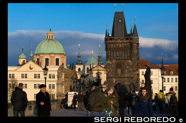 Vistas de la Iglesia de San Nicolás desde el Puente de Carlos.  La Torre de la Iglesia de San Nicolás es una de nuestras preferidas de Praga, la subida es emocionante y tiene varias salas a medio camino que pueden ser visitadas. En estas salas aprenderéis mucho sobre la historia de las campanas y sus diversas funciones. Igual que el Castillo de Praga, las torres del Puente de Carlos y el Ayuntamiento de la Ciudad Vieja, la iglesia de San Nicolás también forma parte indispensable del panorama de Praga. Una de las construcciones barrocas más bellas de Europa corona el barrio Malá Strana desde hace más de tres siglos, siendo el lugar que admira diariamente multitud de personas de todo el mundo. ¡Ríndase usted también ante su belleza y grandiosidad barrocas! La construcción de uno de los símbolos de Praga se debe a la Orden de los jesuitas, que anhelaban edificar su obra más espléndida en Bohemia. Para ello llamaron en busca de ayuda al maestro genial del barroco Kryštof Dientzenhofer. Después de su muerte, continuó su trabajo su hijo, quien proyectó tanto la disposición única de la bóveda como la parte más hermosa de toda la iglesia: la majestuosa cúpula. Más tarde fue construido uno de los campanarios más altos del barrio Malá Strana, surgiendo así una obra sin par al norte de los Alpes en aquella época. Desde los 65 metros de altura que tiene la torre se puede ver todo el barrio de Malá Strana. Durante la época comunista fue utilizada por las fuerzas secretas para vigilar las embajadas extranjeras. 