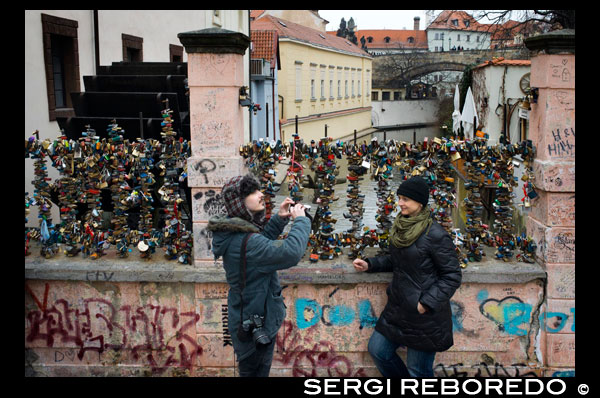 Una pareja en el puente de los enamorados. Praga. No es tan ostentoso ni conocido como el gran Puente de Carlos, pero a los habitantes de Praga les encanta este pequeño y coqueto puente enrejado, casi camuflado en la parte menos conocida del precioso casco antiguo y que con solo 20 metros es capaz de cruzar con extrema confianza nada más y nada menos que el Canal del Diablo. El antiguo puente del Priorato, donde todavía hoy puede verse la vieja rueda del molino, cuenta con su propio duende del agua y desde hace unos años es el lugar más buscado por los enamorados para cerrar sus candados en señar de amor eterno.