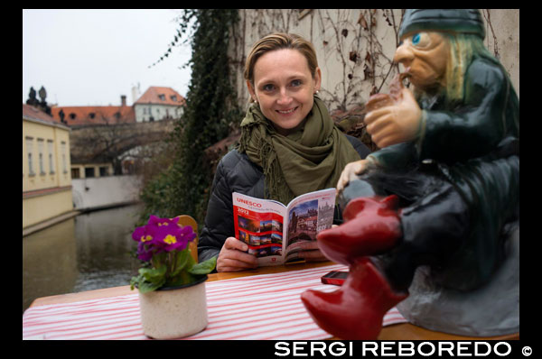 Cafetería junto al puente de los enamorados.  El puente de los enamorados (el antiguo puente del molino del Gran Prior) es el más antiguo que atraviesa el canal del Diablo en Praga. No es muy grande, tiene solo 20,5 m de longitud y 4,3 m de ancho. Através de su rejita se puede ver todavía la rueda del antiguo molino y una estatua de "vodník". Vodník o sea el hombre del agua es típico personaje de los cuentos checos de ni?os en los que representa el mal igual que las brujas. Pero la vista hacia la rueda y el vodník cada vez es más difícil por la cantitad de candados que las personas vienen poniendo en la reja. A ¿qué se debe esta acción? Esta tradición es originaria de China donde al lado de la Gran Muralla China se ponían cuerdas en las que los recién casados solían poner candados cuyas llaves después tiraban através de la muralla para asegurarse el amor eterno y la felicidad. En Europa se celebra esta tradición más gracias al libro "Te quiero" de Federico Moccii. En el libro aparece una pareja que confirma su amor poniendo un peque?o candado en el puente antiguo Milvio en Roma y tirando las llaves al río Tibera. El puente fue elegido a propósito pues en él murió el mártir y patrono de todos los enamorados San Valentin. El libro se ha convertido en un hit, la gente empezó a poner los candados en muchos puentes en toda Europa. En Praga esta costumbre es relativamente nueva. Empezó recién en el a?o 2008. Pero si todavía no han estado, no tengan miedo, su candado todavía cabe. Markéta Lehecková. 