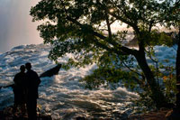 Local tourists visit the Victoria Falls. The Victoria Falls constitutes one of the most spectacular natural wonders of the world. The Local people call it "Mosi-oa-Tunya" -- the smoke that thunders and the Falls are remarkable.  There is a magic about them manifested in the towering column of spray when the river is high, the thunder of the falling water, the terrifying abyss and tranquil lagoons upstream in which hippo and deadly crocodiles lurk. The Victoria falls is 1 708 meters wide, making it the largest curtain of water in the world. It drops between 90m and 107m into the Zambezi Gorge and an average of 550,000 cubic metres of water plummet over the edge every minute. Remarkably preserved in its natural state, Victoria falls inspires visitors as much today as it did David Livingstone in the 1860's. The falls and the surrounding area have been declared National Parks and a World Heritage Site, thus preserving the area from excessive commercialisation. 