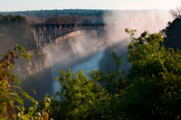 Las vistas de las Cataratas Victoria . Detrás , el puente entre Zambia y Zimbabwe. Durante más de 50 años, el puente fue cruzado con regularidad por los trenes de pasajeros como parte de la ruta principal entre la entonces Rhodesia del Norte , África del Sur y Europa. Los trenes de carga realizan principalmente mineral de cobre ( más tarde, lingotes de cobre ) y madera de Zambia , y el carbón en el país. La edad de los problemas de puentes y mantenimiento han llevado a restricciones de tráfico en horas . Los trenes se cruzan en menos de caminar ritmo y camiones fueron limitados a 30 t , necesitando camiones más pesados ??que hacer un largo desvío a través de la Kazungula Ferry o puente Chirundu . El límite se elevó después de las reparaciones en 2006, pero más fundamental la rehabilitación o construcción de un nuevo puente ha sido ventilado . Durante la crisis de Rhodesian UDI y la guerra de Bush , el puente fue cerrado con frecuencia ( y los servicios regulares de pasajeros no se han reanudado con éxito ) . En 1975 , el puente fue el escenario de las conversaciones de paz fallidos cuando las partes se reunieron en un vagón de tren suspendido sobre el barranco de nueve horas y media . En 1980 los servicios de transporte de mercancías por carretera y se reanudaron y han continuado sin interrupción , excepto para el mantenimiento.