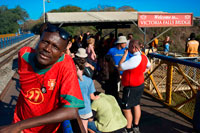 La gente alrededor de Victoria Falls Bridge. The Victoria Falls Puente cruza el río Zambezi justo debajo de las Cataratas Victoria y está construido sobre la Segunda Quebrada de las caídas. A medida que el río es la frontera entre Zimbabwe y Zambia, el puente une los dos países y cuenta con puestos fronterizos en los accesos a los dos extremos, en las ciudades de Victoria Falls, Zimbabwe y Livingstone, Zambia. Durante más de 50 años, el puente fue cruzado con regularidad por los trenes de pasajeros como parte de la ruta principal entre la entonces Rhodesia del Norte, África del Sur y Europa. Los trenes de carga realizan principalmente mineral de cobre (más tarde, lingotes de cobre) y madera de Zambia, y el carbón en el país. La edad de los problemas de puentes y mantenimiento han llevado a restricciones de tráfico en horas. Los trenes se cruzan en menos de caminar ritmo y camiones fueron limitados a 30 t, necesitando camiones más pesados ??que hacer un largo desvío a través de la Kazungula Ferry o puente Chirundu.