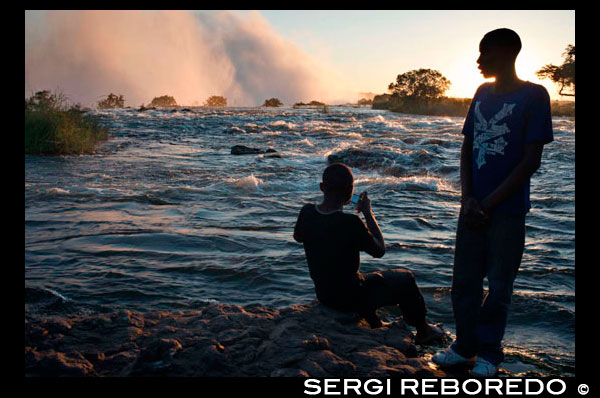 Turistas nacionales visitan las Cataratas Victoria . Estas son algunas de las cascadas más espectaculares del mundo. El río Zambezi , que es más de 2 km de ancho en este punto, se hunde estrepitosamente por una serie de gargantas de basalto y levanta una bruma iridiscente que se pueden ver más de 20 km. El Mosi-oa-Tunya/Victoria Falls es mayor de situación del mundo de la caída del agua y significativo en todo el mundo por sus características geológicas y geomorfológicas excepcionales y los procesos de formación del suelo activas con gran belleza atribuida a las cataratas , es decir, la pulverización , la niebla y el arco iris. Este establecimiento transfronterizo se extiende sobre 6860 hectáreas y comprende 3.779 hectáreas del Parque Nacional de Mosi -oa- Tunya (Zambia ) , 2.340 hectáreas de Parque Nacional de las Cataratas Victoria (Zimbabwe ), 741 hectáreas de la franja ribereña del Zambezi National Park (Zimbabue ) . Una franja ribereña del Parque Nacional Zambezi extiende 9 km al oeste a lo largo de la margen derecha del Zambeze y las islas en el río son todos dentro del parque en cuanto a la palma y las Islas de Kandahar , con las Cataratas Victoria es una de las principales atracciones. La cascada se encuentra a una altitud de unos 915 m sobre el nivel medio del mar ( msnm ) y se extiende hasta alrededor de 1708 m de ancho, con una profundidad media de 100 m, y el punto más profundo de ser 108 m . Sprays de esta cascada gigante se puede ver desde una distancia de 30 km de la carretera de Lusaka , Zambia , y 50 km de camino de Bulawayo , Zimbabwe. Basaltos se han reducido en un sistema fluvial produciendo una serie de gargantas eightspectacular que sirven como criaderos de cuatro especies de aves en peligro de extinción . Los basaltos de la propiedad Victoria Falls Patrimonio Mundial se acodan a diferencia de las del sitio de Patrimonio Mundial de la Calzada de los Gigantes , que son verticales y columnar.